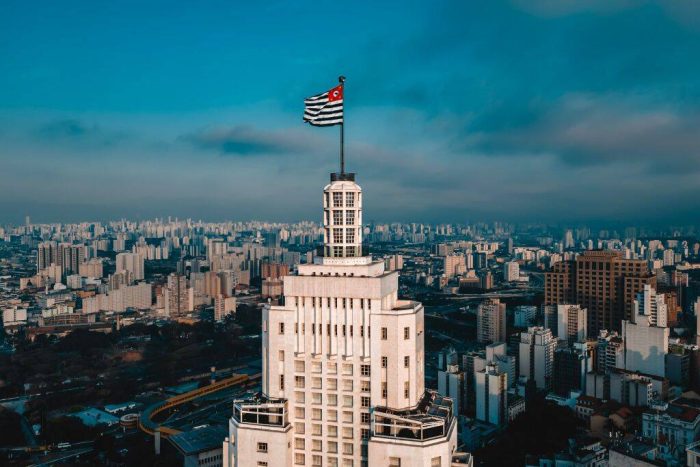 Prédio histórico de São Paulo com a bandeira paulista no topo e vista panorâmica da cidade, no contexto da comparação entre Brasília ou São Paulo.