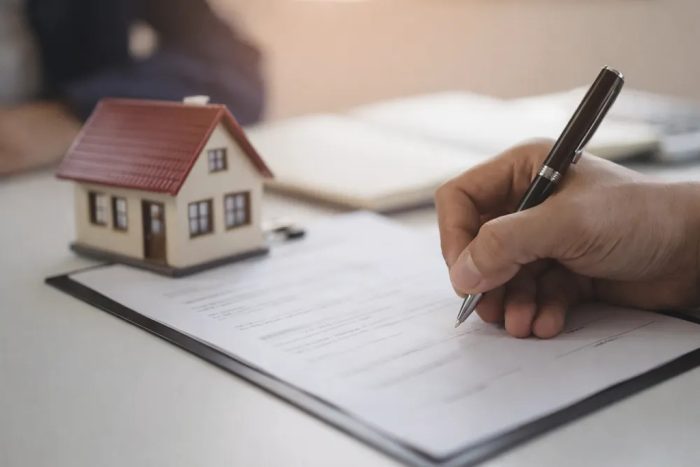 Close de uma pessoa assinando documentos contratuais sobre uma prancheta, ao lado de uma miniatura de casa, representando o processo de como registrar um imóvel pela primeira vez.