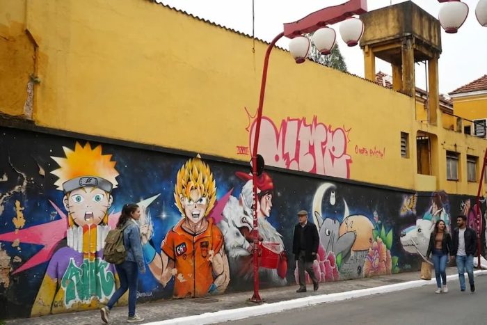 Mural de animes como Naruto e Goku em rua do bairro japonês em São Paulo, com pedestres caminhando ao lado.