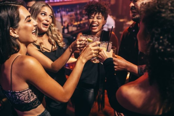 Grupo diverso em ambiente com iluminação mais baixa em um dos bares na Vila Olímpia brinda com drinks enquanto sorriem.